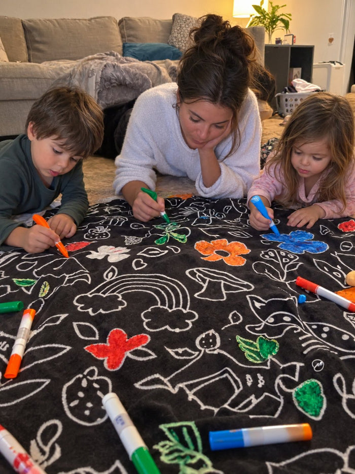 A parent and two children coloring the blanket spread out on the rug